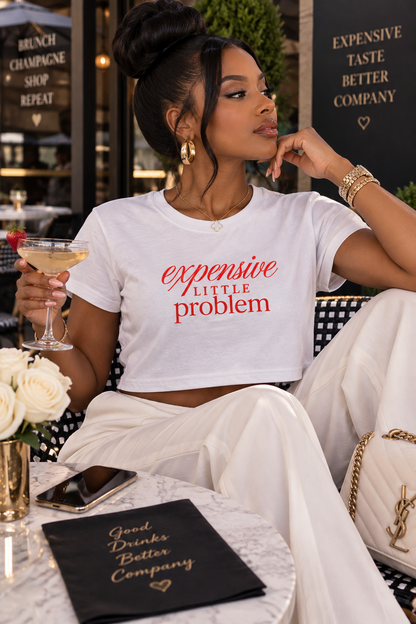 A woman sitting at an outdoor cafe wearing a white crop top t-shirt with the red text 'expensive LITTLE problem' and white wide-leg pants. She is holding a cocktail glass and has a black notebook on a marble table in front of her.