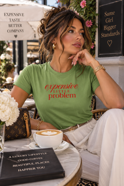 A woman sitting at a cafe table wearing a green graphic t-shirt with the text 'expensive LITTLE problem' in red. She is wearing white pants and holding a white coffee cup.